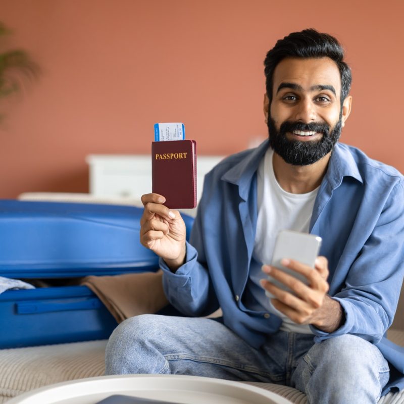 Tickets Booking Service. Cheerful Indian Traveler Man With Phone Showing His Passport With Boarding Pass At Home, Booking And Planning His Vacation Online Via New Travel Application