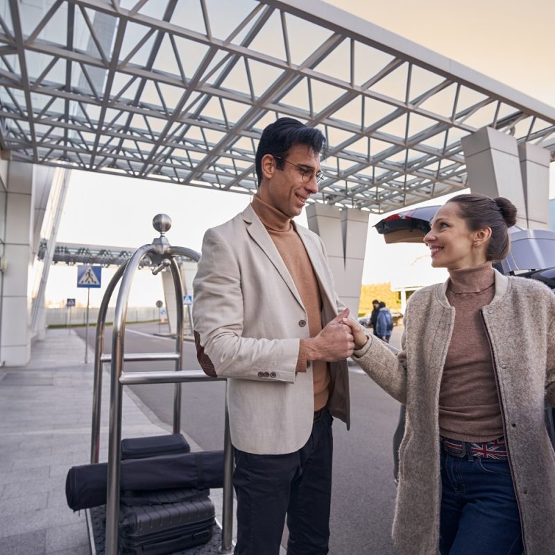 Man standing outside near baggage trolley and holding woman by hand while she exiting car