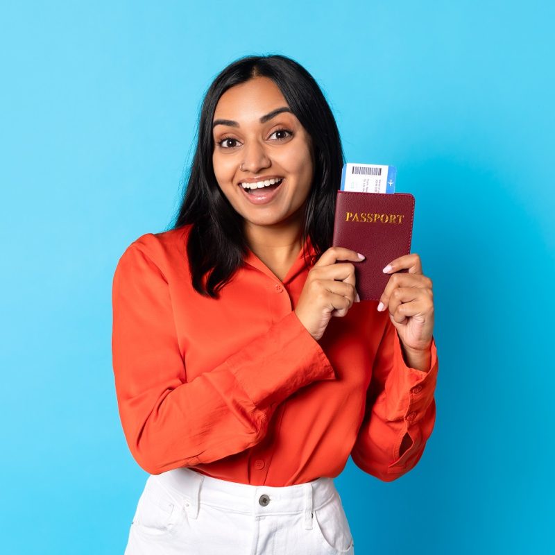 Happy young indian lady traveler showing passport and boarding pass in blue studio, smiling to camera and promoting travel offer for millennials, modern holiday advertisement banner