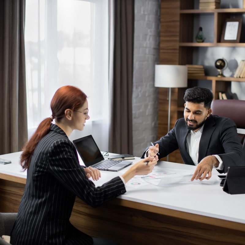 Handsome bearded businessman dealer showing papers with new agreement to his red hair businesswoman customer in a desktop, sitting at table in modern office.