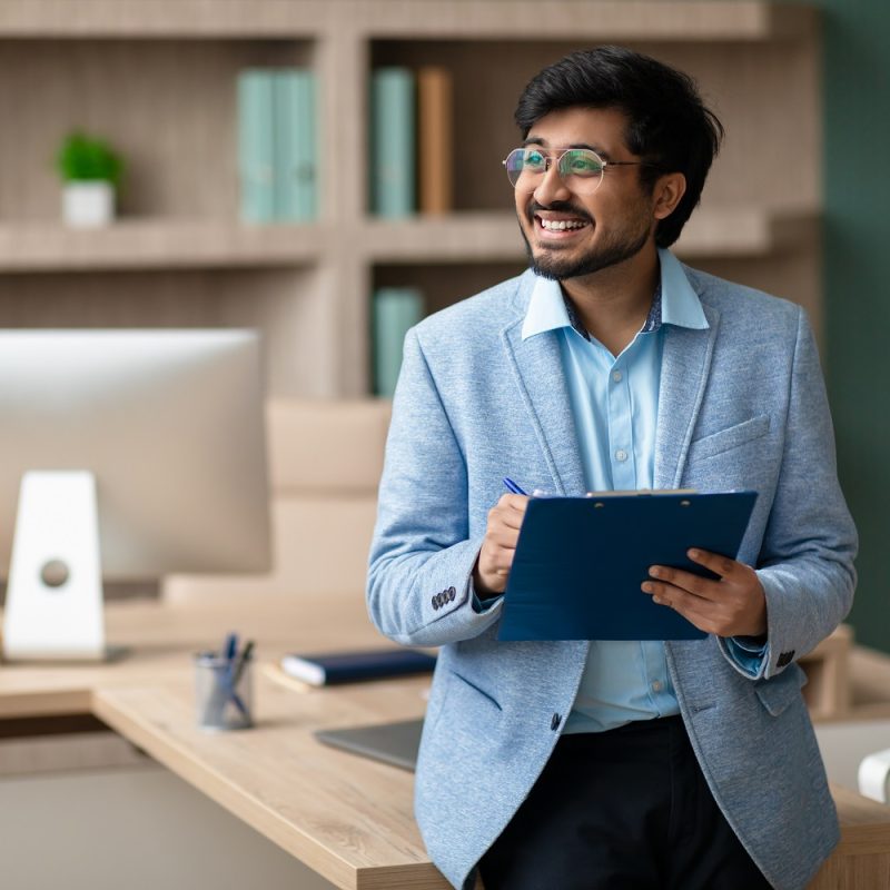 Cheerful Indian professional businessman holds clipboard and signing documents, celebrating successful business deals, standing in modern corporate office interior. Copy space