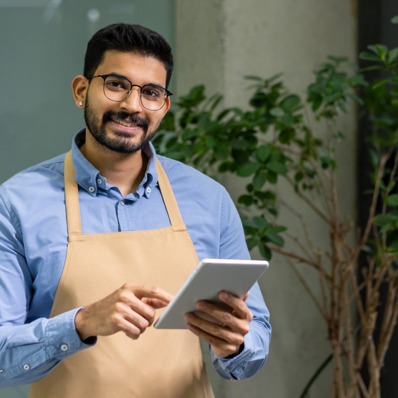 Cheerful male small business owner in apron operates tablet in green plant-filled contemporary workspace, exemplifying modern entrepreneurship.